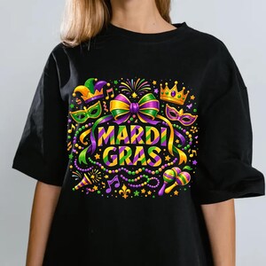 May include: Black t-shirt with a colorful Mardi Gras graphic. The design includes a bow, crown, mask, and the words "Mardi Gras" in a vibrant font. Additional elements include musical notes, beads, and a maraca.