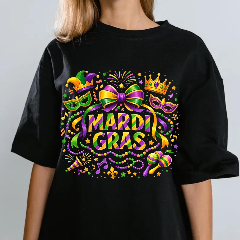 May include: Black t-shirt with a colorful Mardi Gras graphic. The design includes a bow, crown, mask, and the words "Mardi Gras" in a vibrant font. Additional elements include musical notes, beads, and a maraca.