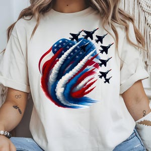May include: Off-white t-shirt with a patriotic design. The graphic depicts a stylized American flag with red, white, and blue smoke trails, and a formation of black fighter jets with red details. The design is centrally placed on the front.