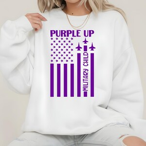 May include: White sweatshirt with a purple graphic. The graphic features a stylized American flag with purple stripes and stars, the words "PURPLE UP" above, and "MILITARY CHILD" in a vertical column. Three purple airplane silhouettes are also included.