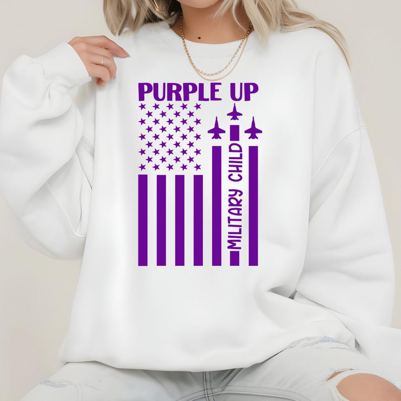 May include: White sweatshirt with a purple graphic. The graphic features a stylized American flag with purple stripes and stars, the words "PURPLE UP" above, and "MILITARY CHILD" in a vertical column. Three purple airplane silhouettes are also included.