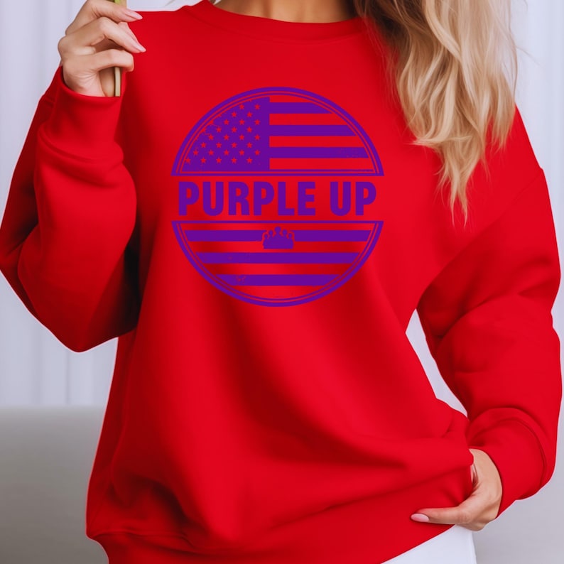 May include: A red sweatshirt with a purple graphic. The graphic is a circle with a purple American flag design and the words "PURPLE UP" in purple letters. The sweatshirt has long sleeves.