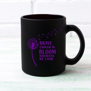 May include: Black ceramic mug with a purple dandelion design and the words "BRAVE ENOUGH TO BLOOM WHEREVER WE LAND." The mug has a curved handle and a smooth, matte finish. The design features a dandelion with seeds blowing away.