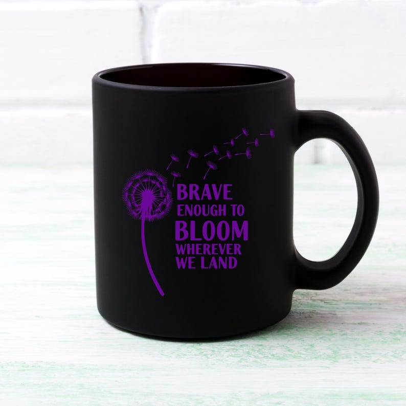May include: Black ceramic mug with a purple dandelion design and the words "BRAVE ENOUGH TO BLOOM WHEREVER WE LAND." The mug has a curved handle and a smooth, matte finish. The design features a dandelion with seeds blowing away.