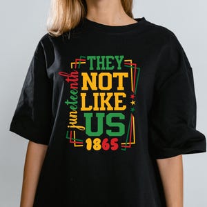 Puede incluir: Camiseta negra con un diseño gráfico colorido. El diseño incluye las palabras "THEY NOT LIKE US 1865" en verde y amarillo, con la palabra "Juneteenth" escrita verticalmente en rojo y amarillo.