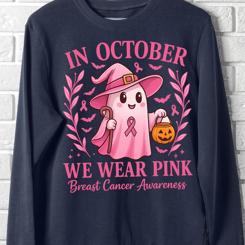 May include: Navy blue sweatshirt with a pink ghost wearing a witch hat, holding a cane and a jack-o'-lantern. Text reads "IN OCTOBER WE WEAR PINK Breast Cancer Awareness". Bats and pink ribbons decorate the design.