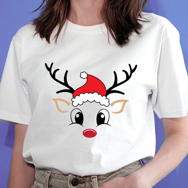 May include: White t-shirt featuring a cartoon reindeer face with black antlers, large eyes, and a red nose. The reindeer wears a red Santa hat with white trim. A festive design for the holiday season.