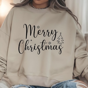 May include: Beige sweatshirt with the words "Merry Christmas" in black script. The design includes snowflakes and a Christmas tree. The sweatshirt has a crew neck and long sleeves.