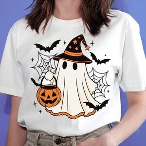 May include: A white short-sleeved t-shirt featuring a graphic of a white ghost wearing a black witch hat with orange stars. The ghost holds an orange jack-o'-lantern bucket, surrounded by black spiderwebs, flying bats, and small star accents.