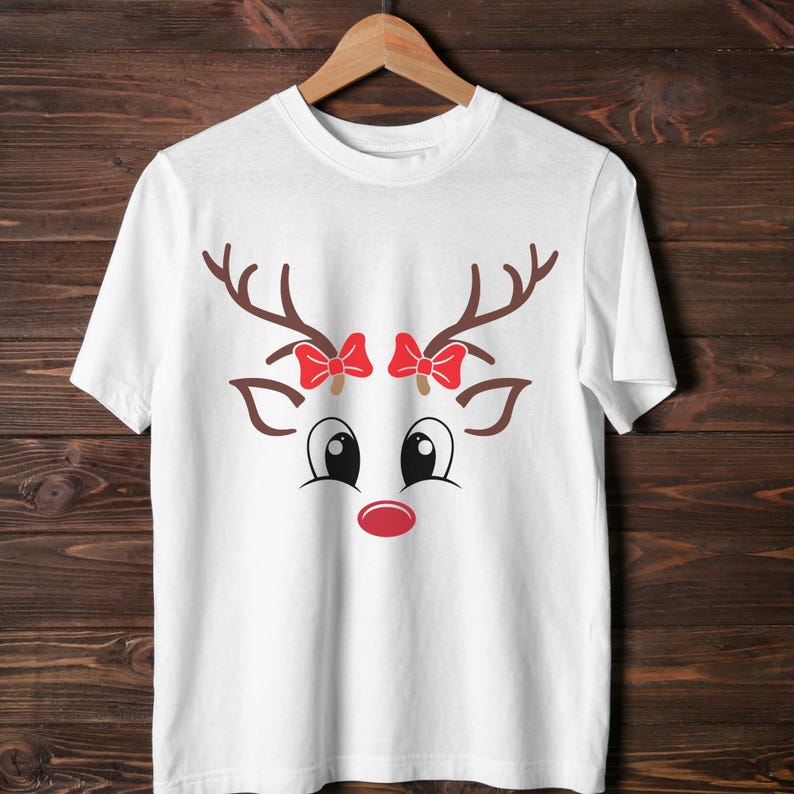 May include: White t-shirt featuring a reindeer face design. The design includes brown antlers, red bows, black eyes, and a red nose. The shirt is hanging on a wooden hanger.