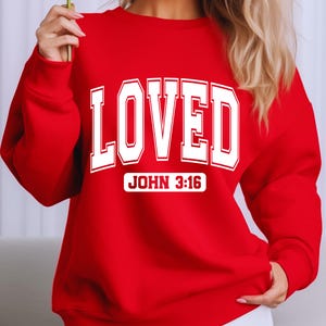 May include: A red sweatshirt with the word "LOVED" in large white block letters. Below the word "LOVED" is the text "JOHN 3:16" in a white rectangular box. The sweatshirt is a solid red color.