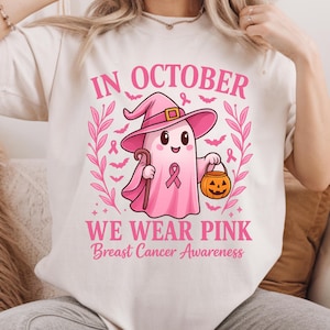 May include: Cream-colored t-shirt with a pink graphic. The graphic features a cartoon ghost wearing a witch hat and holding a cane and a jack-o'-lantern. Text reads "IN OCTOBER WE WEAR PINK Breast Cancer Awareness."