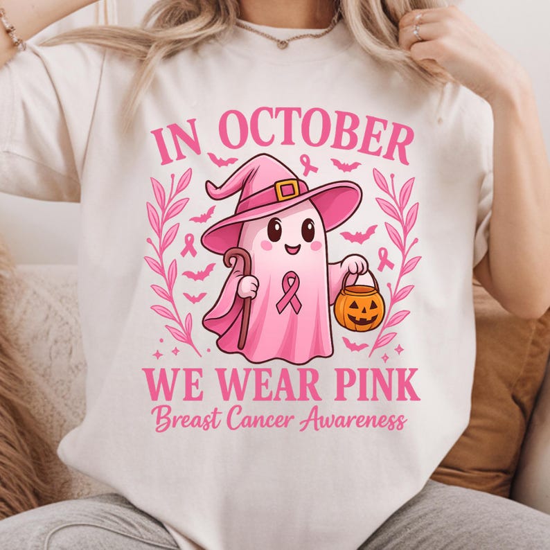 May include: Cream-colored t-shirt with a pink graphic. The graphic features a cartoon ghost wearing a witch hat and holding a cane and a jack-o'-lantern. Text reads "IN OCTOBER WE WEAR PINK Breast Cancer Awareness."