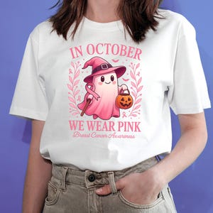 May include: White t-shirt with a pink graphic. The graphic features a ghost wearing a witch hat and holding a pumpkin. Text on the shirt reads "IN OCTOBER WE WEAR PINK Breast Cancer Awareness".