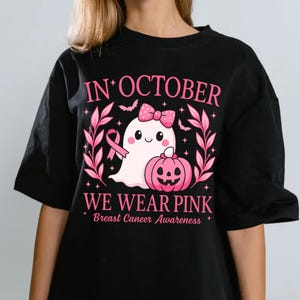 May include: Black t-shirt with a pink graphic for Breast Cancer Awareness. The design features a cute ghost with a pink bow, a pink pumpkin, and the text "IN OCTOBER WE WEAR PINK".
