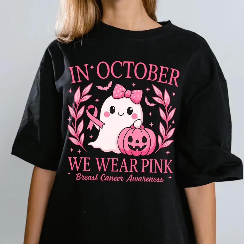 May include: Black t-shirt with a pink graphic for Breast Cancer Awareness. The design features a cute ghost with a pink bow, a pink pumpkin, and the text "IN OCTOBER WE WEAR PINK".