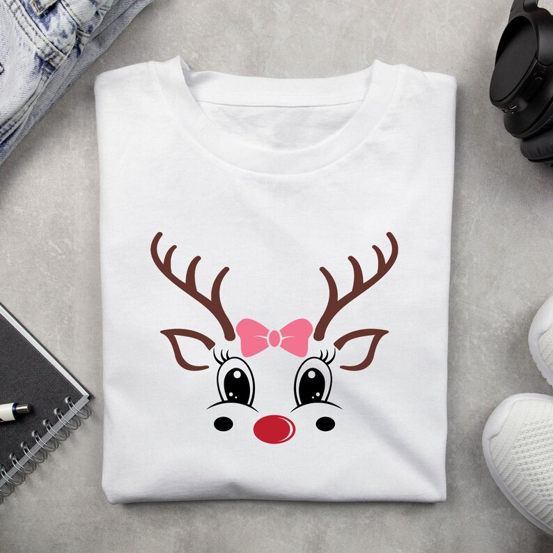 May include: White t-shirt featuring a cartoon reindeer face with brown antlers, large black eyes, a pink bow, and a red nose. The design is centered on the front of the shirt, perfect for holiday or Christmas wear.