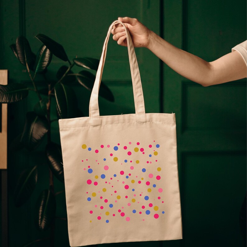 K&ouml;nnte beinhalten: Eine wei&szlig;e Canvas-Tragetasche mit einem bunten Konfetti-Design. Das Konfetti ist in den Farben Pink, Blau und Gold.