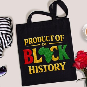 K&ouml;nnte beinhalten: Schwarze Tragetasche mit dem Text "PRODUCT OF BLACK HISTORY" in Gelb, Rot und Gr&uuml;n. Das Design beinhaltet eine Umrisszeichnung des afrikanischen Kontinents. Die Tasche hat schwarze Griffe.
