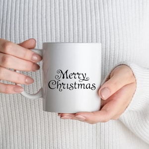 May include: A white ceramic mug with the words "Merry Christmas" in elegant black script. The mug is held by a person wearing a white sweater. The mug has a standard handle and a smooth, glossy finish, perfect for holiday beverages.