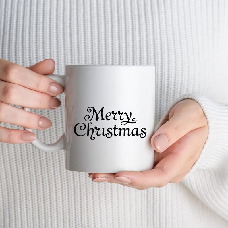 May include: A white ceramic mug with the words "Merry Christmas" in elegant black script. The mug is held by a person wearing a white sweater. The mug has a standard handle and a smooth, glossy finish, perfect for holiday beverages.