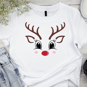 May include: White t-shirt featuring a reindeer face design. The design includes brown antlers, large black eyes with eyelashes, pink cheeks, and a red nose. A festive and playful graphic tee.