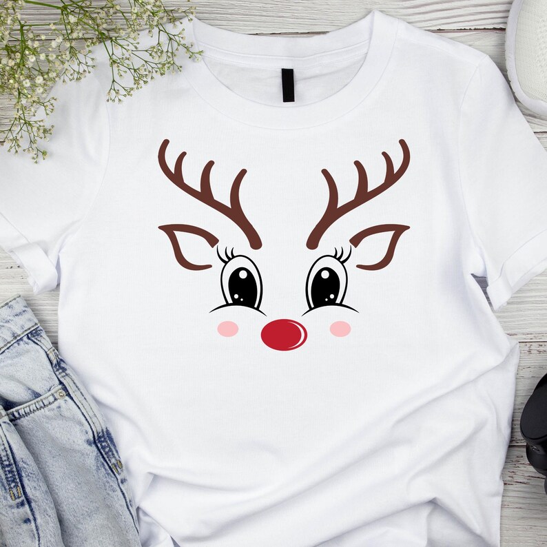 May include: White t-shirt featuring a reindeer face design. The design includes brown antlers, large black eyes with eyelashes, pink cheeks, and a red nose. A festive and playful graphic tee.