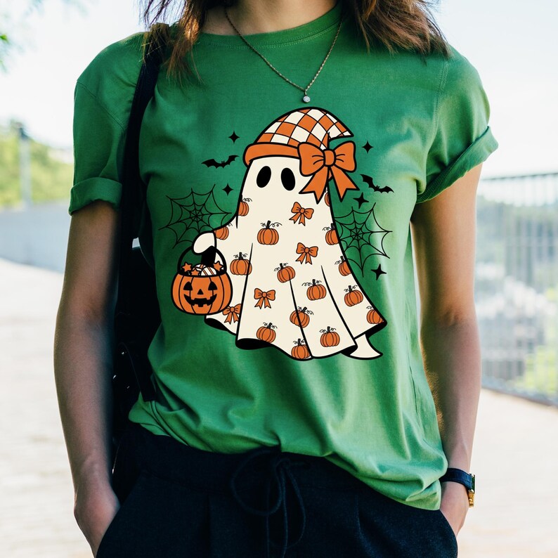 May include: A green short-sleeved t-shirt featuring a graphic of a white ghost wearing an orange and white chequered hat with a bow. The ghost is holding an orange jack-o'-lantern bucket and is surrounded by small pumpkins, bows, and spiderwebs.