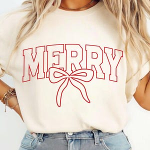 Könnte beinhalten: Cremefarbenes T-Shirt mit dem Wort "MERRY" in roten Blockbuchstaben und einer roten Schleifen-Grafik. Das Shirt ist ein lässiger Stil, passend für festliche Anlässe.
