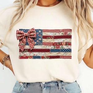 May include: Cream-colored t-shirt featuring a patchwork American flag design. The flag incorporates red, white, and blue fabrics with floral and star patterns, and a plaid bow. A patriotic and stylish apparel item.