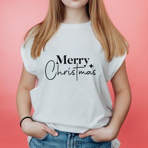 May include: White t-shirt with the words "Merry Christmas" in black script. The shirt has a crew neck and short sleeves. The background is a solid pink color.
