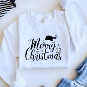 May include: White crew neck sweatshirt with the words "Merry Christmas" in a script font, along with a Santa hat, snowflake, and Christmas tree graphic in black. The sweatshirt is folded on a white surface.