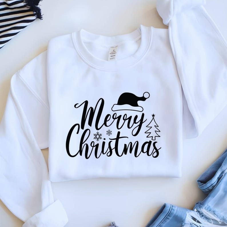 May include: White crew neck sweatshirt with the words "Merry Christmas" in a script font, along with a Santa hat, snowflake, and Christmas tree graphic in black. The sweatshirt is folded on a white surface.