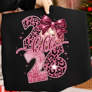 Pode incluir: Camiseta preta com um design de glitter rosa. O design inclui os números "26", a palavra "hello", uma bola de discoteca, um laço e estrelas. Os números têm um padrão de leopardo.