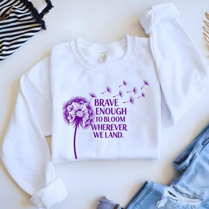 May include: White sweatshirt with a purple dandelion graphic and the text "BRAVE ENOUGH TO BLOOM WHEREVER WE LAND." The design features a detailed dandelion with seeds blowing away. The sweatshirt has long sleeves and a crew neck.