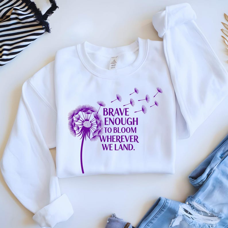 May include: White sweatshirt with a purple dandelion graphic and the text "BRAVE ENOUGH TO BLOOM WHEREVER WE LAND." The design features a detailed dandelion with seeds blowing away. The sweatshirt has long sleeves and a crew neck.