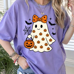 May include: A light purple t-shirt featuring a graphic of a white ghost wearing an orange bow with black stars. The ghost is holding an orange jack-o'-lantern bucket and is surrounded by small orange pumpkins, black bats, and a spider web.