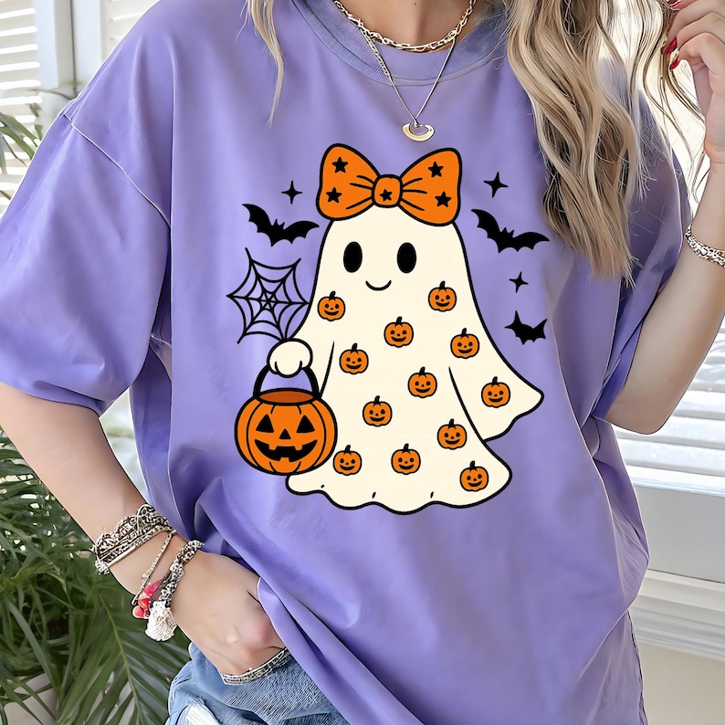 May include: A light purple t-shirt featuring a graphic of a white ghost wearing an orange bow with black stars. The ghost is holding an orange jack-o'-lantern bucket and is surrounded by small orange pumpkins, black bats, and a spider web.