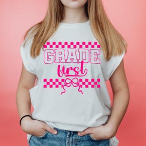 Op de afbeelding: Wit T-shirt met de roze tekst "GRADE first", met een geblokt patroon erboven en eronder. Het T-shirt heeft een strik ontwerp en wordt door een persoon gedragen.