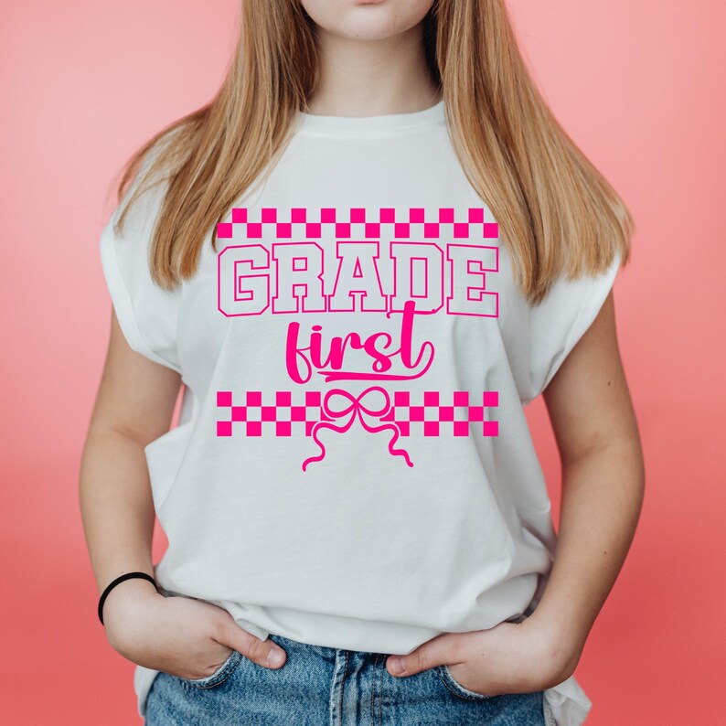 Op de afbeelding: Wit T-shirt met de roze tekst "GRADE first", met een geblokt patroon erboven en eronder. Het T-shirt heeft een strik ontwerp en wordt door een persoon gedragen.