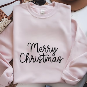 May include: A light pink sweatshirt with the words "Merry Christmas" in black script. The crew neck sweatshirt is folded, with a portion of the sleeve visible. The background includes a straw bag, sunglasses, and a brown hat.