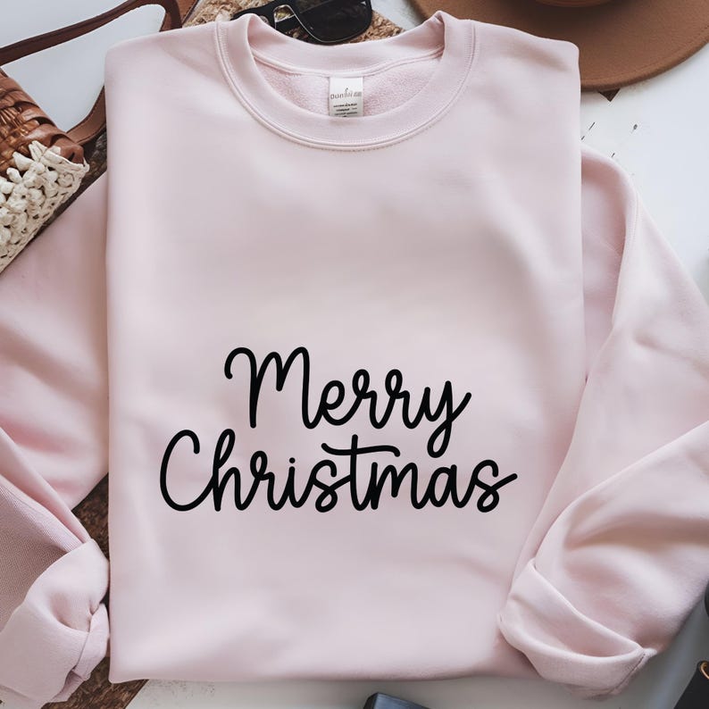 May include: A light pink sweatshirt with the words "Merry Christmas" in black script. The crew neck sweatshirt is folded, with a portion of the sleeve visible. The background includes a straw bag, sunglasses, and a brown hat.