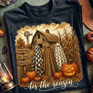May include: A dark grey t-shirt featuring a graphic of a rustic barn, a windmill, and corn stalks. Three ghosts with checkered, leopard, and polka dot patterns stand in front. A sign reads PUMPKINS, HAY RIDES, CORN MAZE. Text reads tis the season.