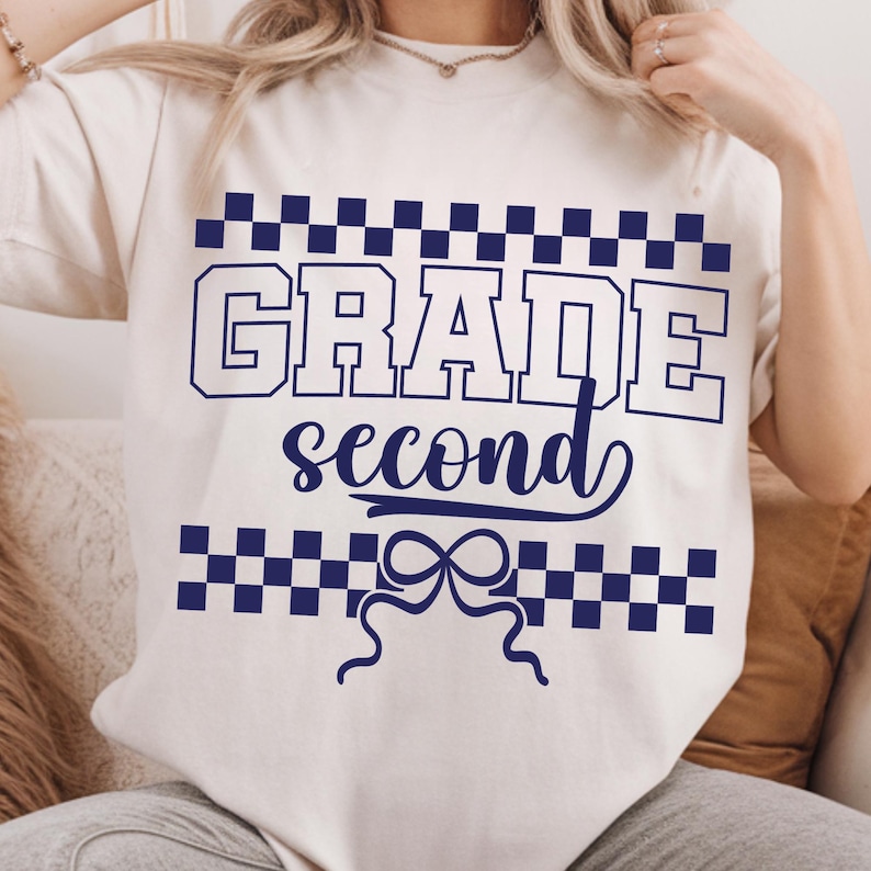 Op de afbeelding: Cr&egrave;mekleurig T-shirt met de woorden "GRADE second" in marineblauw. De tekst is omlijst door een dambordpatroon en een decoratieve strik. Het shirt is een casual stijl.