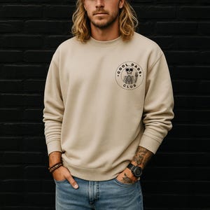 May include: A beige crewneck sweatshirt with a circular graphic on the chest. The graphic features a skeleton wearing sunglasses and the words "COOL DADS CLUB". The sweatshirt is a neutral color and has a classic, casual style.