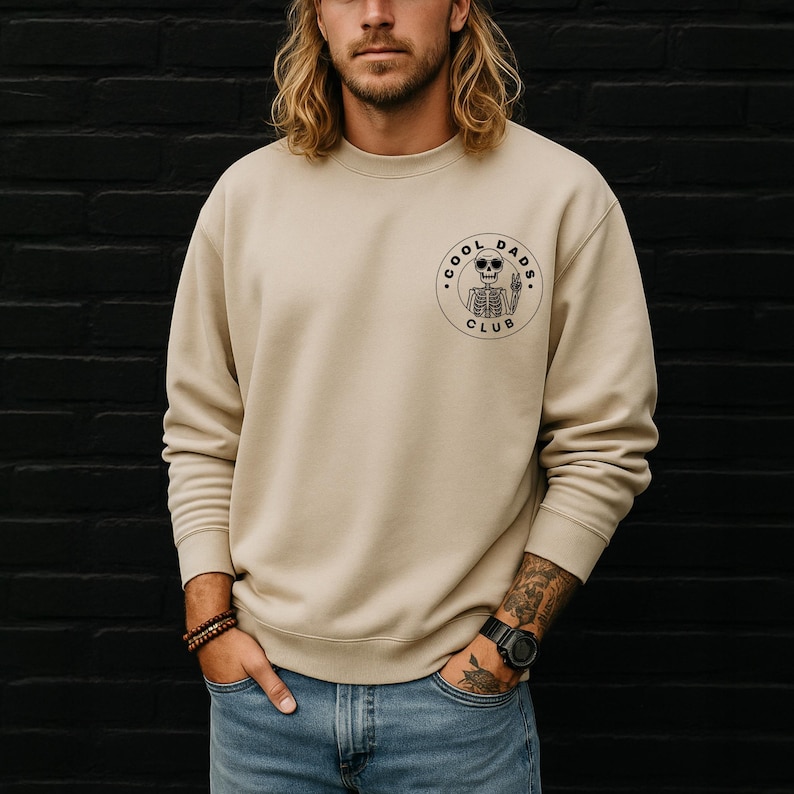 May include: A beige crewneck sweatshirt with a circular graphic on the chest. The graphic features a skeleton wearing sunglasses and the words "COOL DADS CLUB". The sweatshirt is a neutral color and has a classic, casual style.