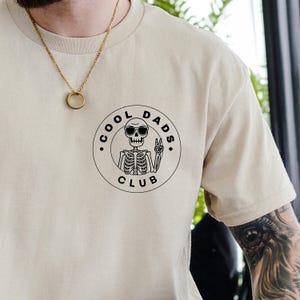 May include: Beige t-shirt with a black graphic of a skeleton wearing sunglasses and giving a peace sign, encircled by the words "COOL DADS CLUB." The shirt has a crew neck and short sleeves. A gold necklace is visible.