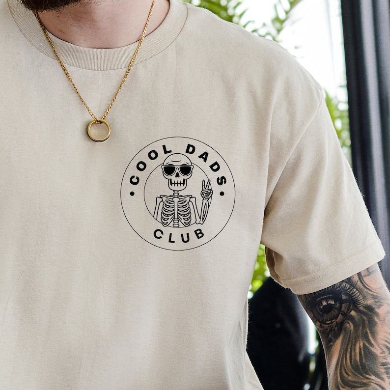 May include: Beige t-shirt with a black graphic of a skeleton wearing sunglasses and giving a peace sign, encircled by the words "COOL DADS CLUB." The shirt has a crew neck and short sleeves. A gold necklace is visible.