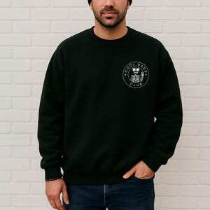 May include: Dark green sweatshirt with a crew neck. The sweatshirt features a white circular graphic on the left chest that reads "COOL DADS CLUB" around a skeleton holding a beer. The person is wearing blue jeans.