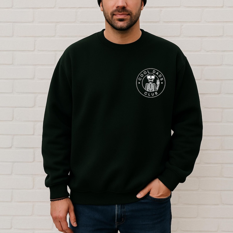 May include: Dark green sweatshirt with a crew neck. The sweatshirt features a white circular graphic on the left chest that reads "COOL DADS CLUB" around a skeleton holding a beer. The person is wearing blue jeans.